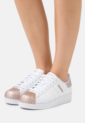 Hvide platform-sneakers med metalliske rosaguld-tåkapper og matchende hælstropper, båret på bare fødder mod en ensfarvet baggrund.