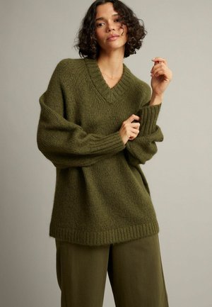 Vrouw met krullend haar die een oversized olijfgroene gebreide trui en bijpassende losse broek draagt tegen een effen achtergrond.