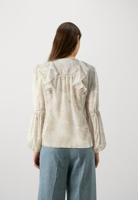 Femme aux longs cheveux bruns portant un chemisier crème à motif paisley avec des volants et des manches bouffantes, associé à un jean large bleu clair, de dos.