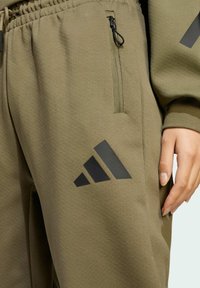 Olivové tepláky vyrobené z mäkkej tkaniny, s vreckom na zips a čiernym logom Adidas na ľavom stehne.