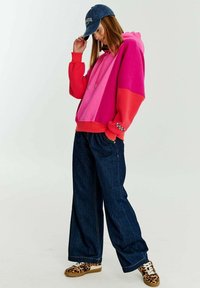 Mikina s kapucí ve dvoubarevném provedení v červené, růžové a magentové barvě, v kombinaci s džínami s širokými nohavicemi a teniskami s leopardím potiskem.