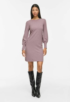 Femme portant une robe en maille mauve à manches longues bouffantes et des bottes en cuir noires montantes jusqu'aux genoux, debout devant un fond blanc.