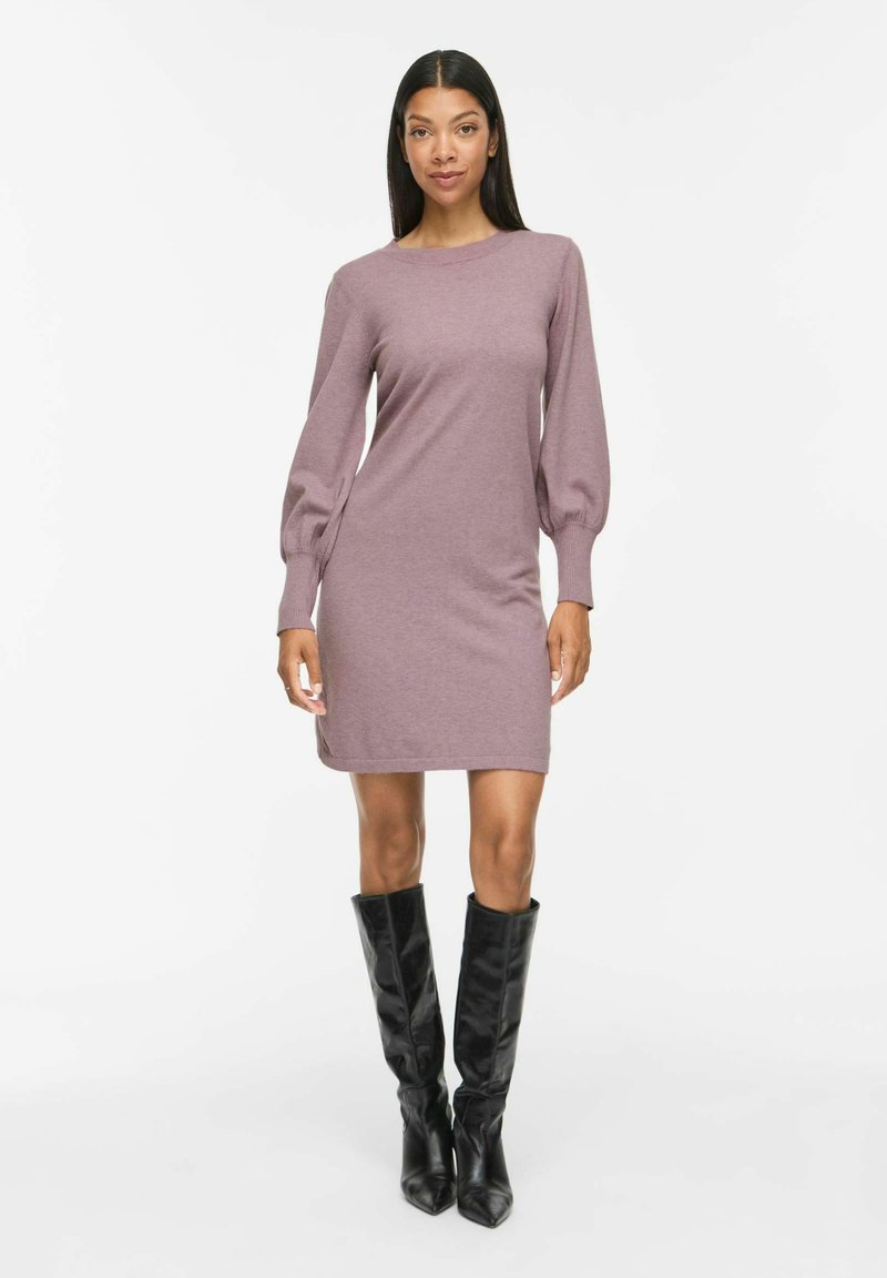 Femme portant une robe en maille mauve à manches longues bouffantes et des bottes en cuir noires montantes jusqu'aux genoux, debout devant un fond blanc.
