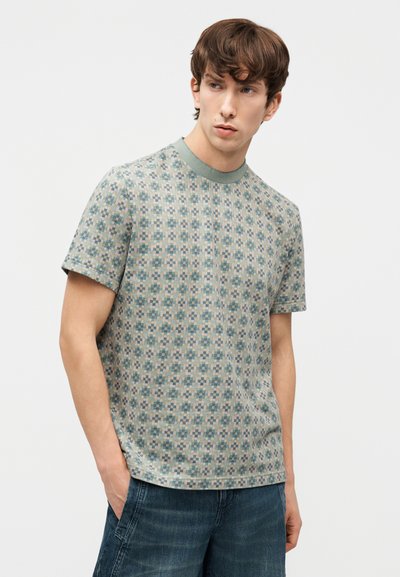 Νεαρός άνδρας με καστανά μαλλιά φοράει κοντομάνικο πουλόβερ με σχέδιο και σκούρο μπλε τζιν, με τα χέρια στις τσέπες, κοιτάζοντας πλάγια.