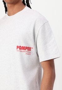 Grey cotton T-shirt featuring a crew neck and short sleeves. Notable red text reads "POMPEII™ We Champion Walking (Madrid)."