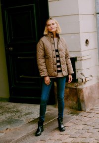 Quilted brown jacket with black buttons, worn over a beige striped sweater, paired with dark skinny jeans and black ankle boots.