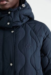 Marineblauwe gewatteerde pufferjack met hoge kraag, zilveren drukkers en geborduurd "Lee" logo. Gladde textuur en golvend ontwerp.