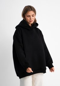 Hoodie oversize nero realizzato in tessuto morbido, caratterizzato da una grande tasca frontale e un cappuccio con cordino. Presenta maniche ampie e polsini a coste.