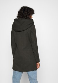 Manteau à capuche vert foncé au design structuré, fabriqué en tissu texturé. Présente une silhouette épurée et des coutures visibles.