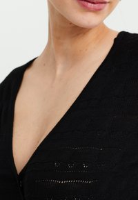 Top en tricot noir avec un décolleté en V profond, un motif texturé et des détails ajourés subtils près du col. Gros plan sur le tissu et le design.