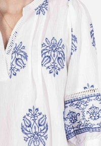Blouse en tissu blanc avec un riche broderie bleue aux motifs floraux. L'encolure dispose d'une patte de boutonnage et les manches sont froncées avec une finition en dentelle.