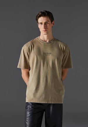 Světle hnědé oversized tričko s kulatým výstřihem, krátkými rukávy a nápisem "Zign Studio Berlin" vytištěným na přední straně tmavým písmem.