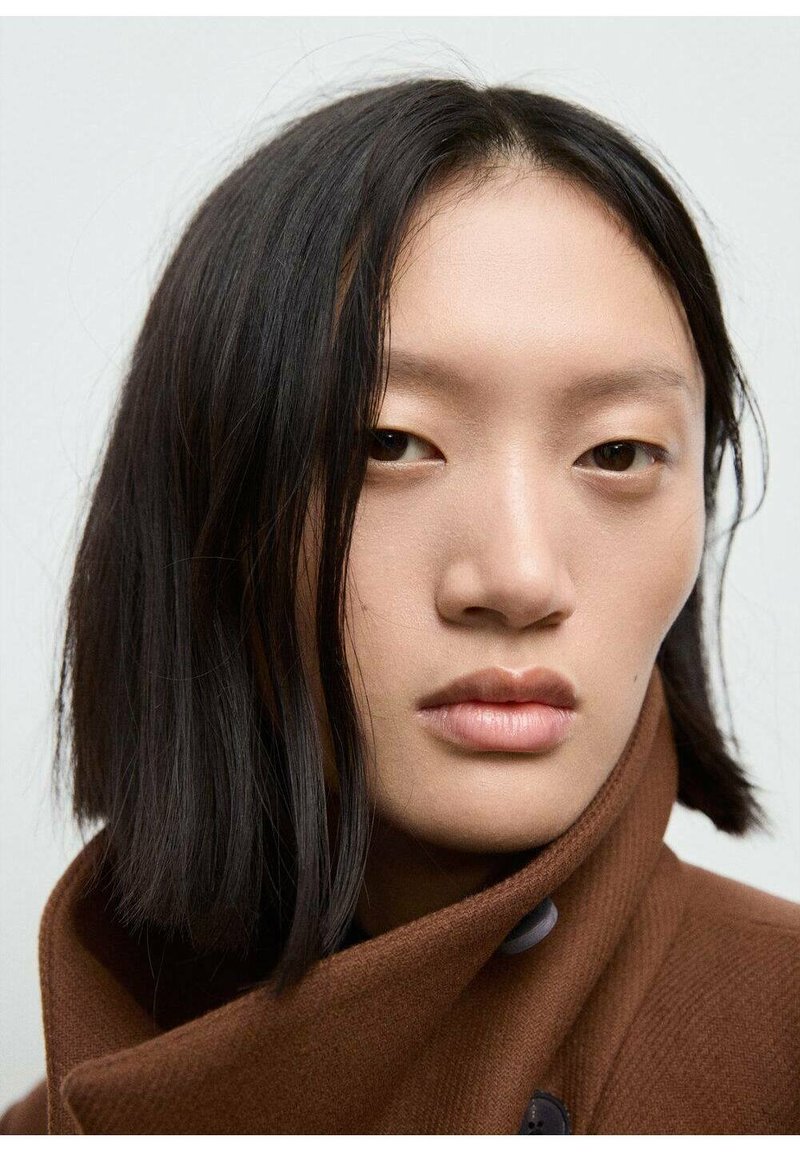 Manteau en laine brun avec un col haut et des boutons, porté par une personne aux cheveux raides et foncés. Fond neutre, accent sur les traits du visage.