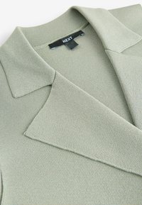 Zelený pletený blazer s formovaným límcem a texturovanou tkaninou, s hladkým vnitřním podšíváním a štítkem s nápisem "NEXT."