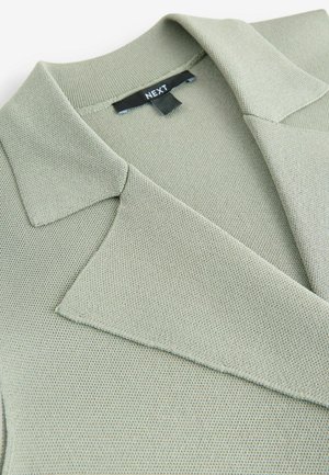 Blazer de punto verde con un cuello estructurado y una tela texturizada, con un forro interior suave y una etiqueta que dice "NEXT."
