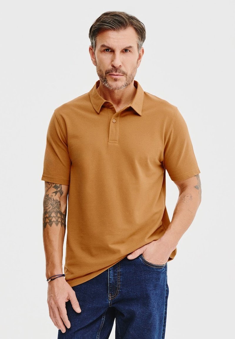 Koszulka polo z krótkim rękawem w brązowym kolorze z klasycznym kołnierzykiem, z dwoma guzikami i gładką teksturą. W połączeniu z niebieskimi dżinsami.