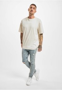 Kremhvitt oversized t-skjorte med korte ermer, kombinert med slitte blå jeans og hvite joggesko. Tatoveringer synlige på armene. Enkel, uformell design.
