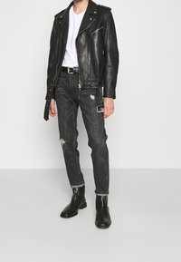 Hombre con chaqueta de cuero negra, camiseta blanca de cuello en V, jeans negros desgastados con rotos, cinturón negro y botas de cuero negras de pie sobre un fondo liso.