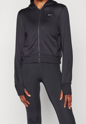 Person iført sort hættetrøje med lynlås, hætte og tommelfingerhuller på ærmerne, kombineret med sorte leggings, står foran en ensfarvet lys baggrund.