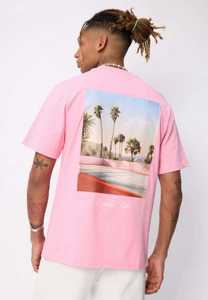 Homme avec des dreadlocks portant un T-shirt rose avec un imprimé de terrain de basket et de palmiers, ainsi qu’un short blanc, face à l’objectif mais de dos.