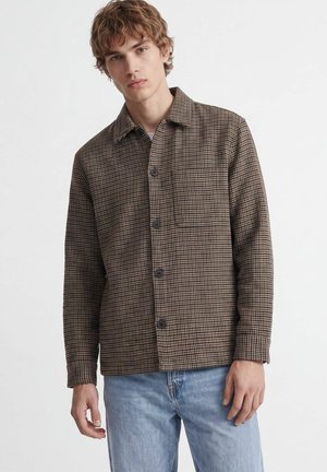Jeune homme portant une veste-chemise à carreaux marron avec une poche avant et un jean bleu clair sur un fond blanc uni.