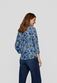 Blusenoberteil mit floralem Druck in Blautönen, mit 3/4-Ärmeln und rundem Ausschnitt. Kombiniert mit dunkelblauen Jeans, die lebendige Muster zeigen.