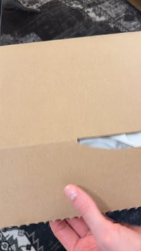 Ręka trzymająca częściowo otwarte, zwykłe kartonowe pudełko z widoczną białą tkaniną w środku, na czarno-białej, wzorzystej powierzchni.