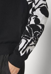 Svart tröja med ett strukturerat svart- och vitt blommönster på vänster ärm; ribbade manschetter och nederkant som visar upp kontrasterande stickade stilar.