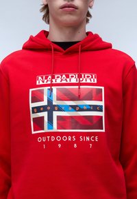 Rode hoodie met een grafisch ontwerp van een gestileerde Noorse vlag, de tekst "Napapuri" en "geographic," met de datum "1987." Zachte stof, regular fit.