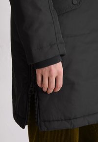 Czarna kurtka puchowa o gładkiej teksturze, z bocznym zapięciem na zamek oraz rękawem z mankietem w okolicy nadgarstka, częściowo widoczna ręka.