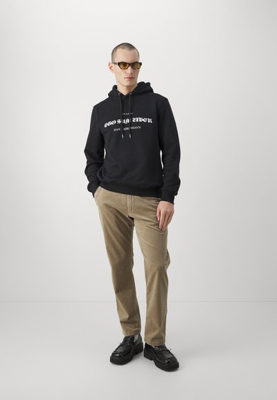 Haut à capuche noir avec texte blanc, pantalon en velours côtelé beige et chaussures noires à semelle épaisse. Le modèle porte des lunettes de soleil teintées jaunes, posé contre un fond neutre.