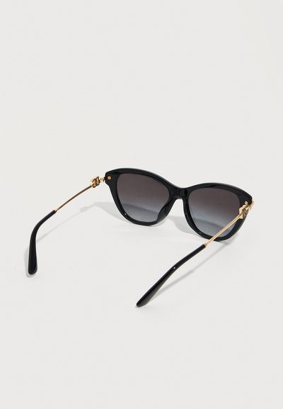 Μαύρα γυαλιά ηλίου με σκελετό γάτας, με φακούς σκοτεινούς με διαβάθμιση και λεπτούς χρυσούς μεταλλικούς βραχίονες που ακουμπούν σε λευκή επιφάνεια.