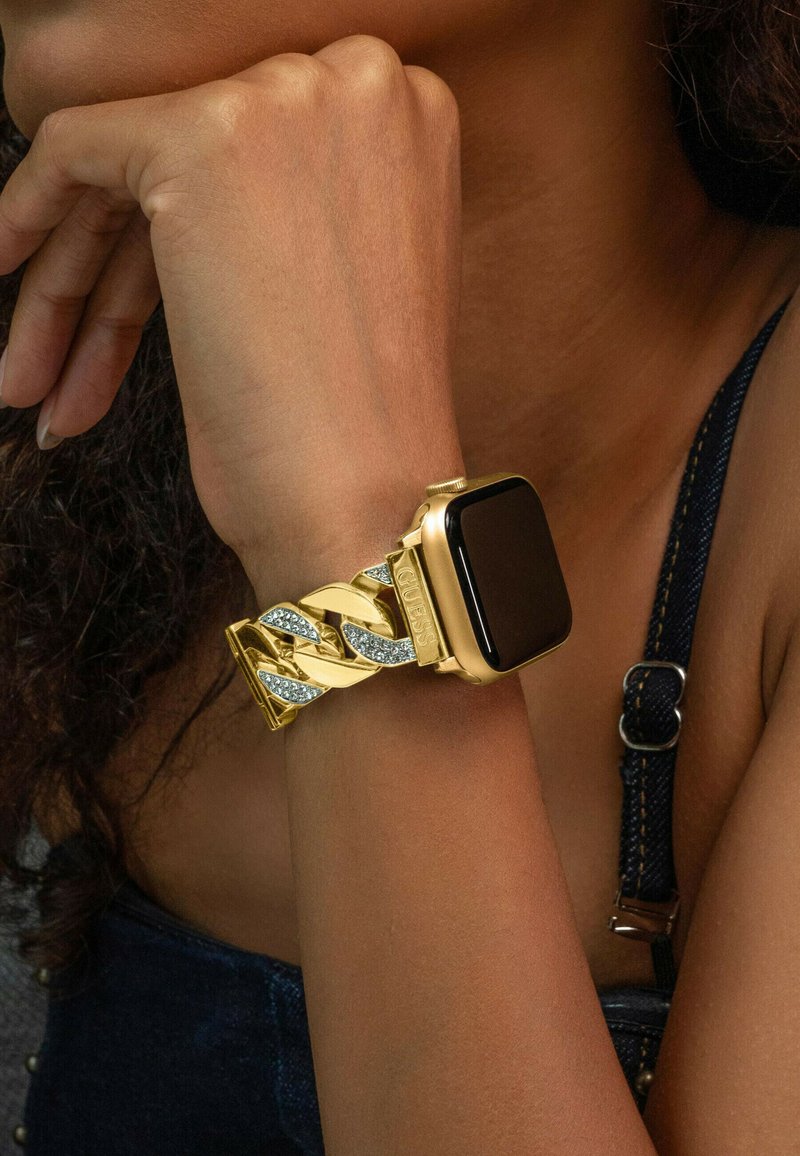 Złoty smartwatch z bransoletą w kształcie ogniwa łańcucha, z błyszczącymi ogniwami i wbudowanymi kryształkami. Czarna kwadratowa tarcza zegarka. Teksturowane tło przypominające skórę.