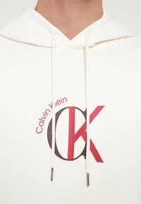 Hanorac alb cu snur. Are un logo grafic în roșu și gri, cu textul "Calvin Klein" și "CK". Textură netedă, design casual.