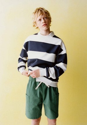 Blondes Kind trägt einen schwarz-weiß gestreiften Pullover und grüne Shorts und zieht an der Kordel der Shorts vor einem gelben Hintergrund.