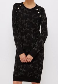 Svart stickad klänning med ett subtilt kamouflage-mönster, prydd med silverfärgade knappar vid axlarna och ribbade ärmslut.