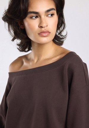 Jonge vrouw met kort donker haar kijkt opzij, draagt een losvallende donkerbruine off-shoulder top tegen een effen lichte achtergrond.