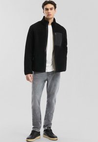 Schwarze Fleecejacke mit hohem Kragen und Reißverschluss-Brusttasche, kombiniert mit grauen Jeans und schwarzen Sneakers. Glatte Textur, lässiges Design.
