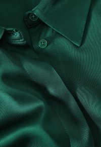 Camicia di raso verde smeraldo con colletto classico, dotata di due bottoni coordinati e texture liscia e lucida. Visibili dettagli di cucitura sottili.
