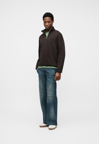 Άνδρας που στέκεται με τα χέρια στις τσέπες, φορώντας μαύρο πουλόβερ με φερμουάρ μέχρι τη μέση, πράσινο πουκάμισο, μπλε τζιν παντελόνι με ευρύ πόδι και άσπρα παπούτσια, σε απλό φόντο.