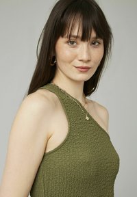 Haut sans manches vert olive, texturé, avec un col haut et un design asymétrique. Porté avec des colliers en or superposés.