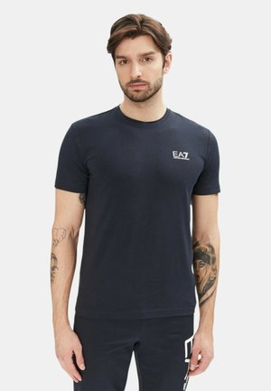 Uomo con capelli castani e barba chiara che indossa una maglietta EA7 blu navy e pantaloni abbinati, mostrando tatuaggi su entrambi gli avambracci su uno sfondo semplice.