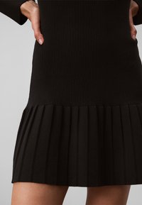 Femme portant une robe en maille côtelée noire avec un ourlet plissé, les mains posées sur les hanches sur un fond neutre.