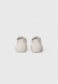 Light beige suede sneakers with a low-top design, rounded back, and rubber soles. Features minimal stitching and a logo at the heel.