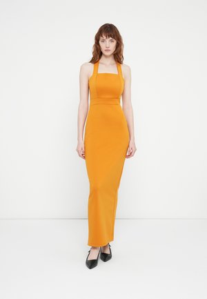 Lang, getailleerde oranje jurk met een vierkante halslijn en halterbandjes, gemaakt van gladde stof, gecombineerd met zwarte puntige hakken.