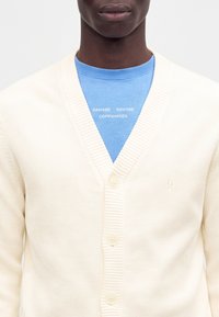 Gros plan d'une personne portant un cardigan en maille crème sur une chemise bleue avec un texte blanc indiquant Samsøe Samsøe Copenhagen.