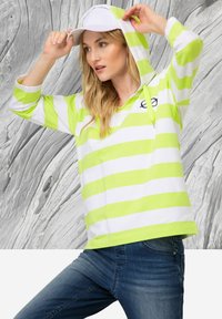 Felpa a righe verde e bianca con tasca a marsupio frontale, cordoncino e dettaglio del logo. Abbinata a jeans blu e un cappellino bianco.