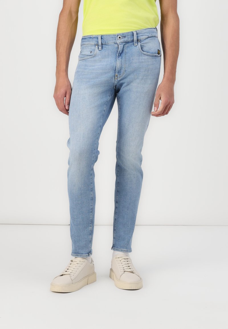 Világoskék, slim fit farmernadrág, öt zsebbelső kialakítással és finom kopással. Bézs sportcipővel tálalva, hogy kontrasztot teremtsen.