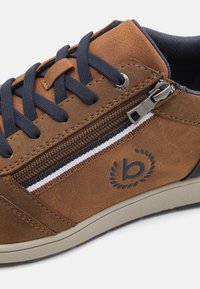 Sneaker din piele ecologică maro cu sireturi bleumarin și buzunar cu fermoar. Are o accentuare cu dungi albe și bleumarin și un logo negru pe lateral.