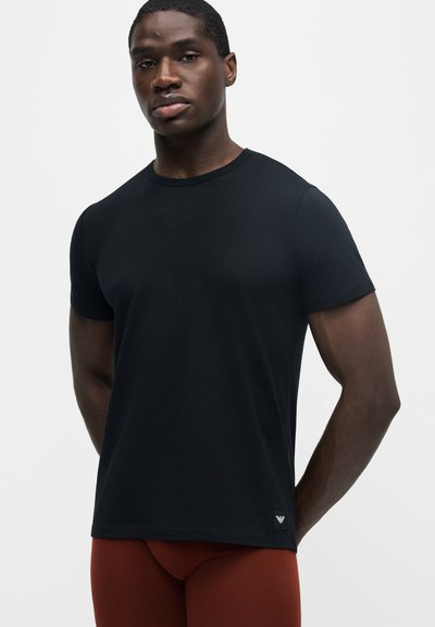 Homme portant un t-shirt noir uni à manches courtes avec un petit logo près de l'ourlet et un pantalon couleur rouille, debout sur un fond blanc.